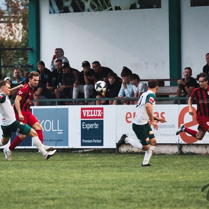 Fußballspieler konkurrieren auf einem Feld, während eine Menge von den Tribünen aus zuschaut. Der Spieler in Weiß kickt den Ball, während der Spieler in Rot versucht, ihn zu blocken.