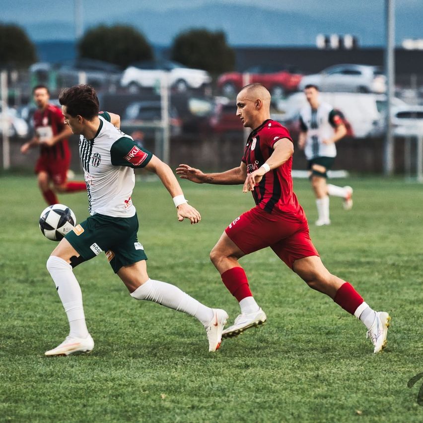 Zwei Fußballspieler sind auf einem Feld in Aktion, einer in einem roten Trikot und der andere in Grün. Der Spieler in Rot jagt den Ball, während der Spieler in Grün versucht, ihn zu treten.