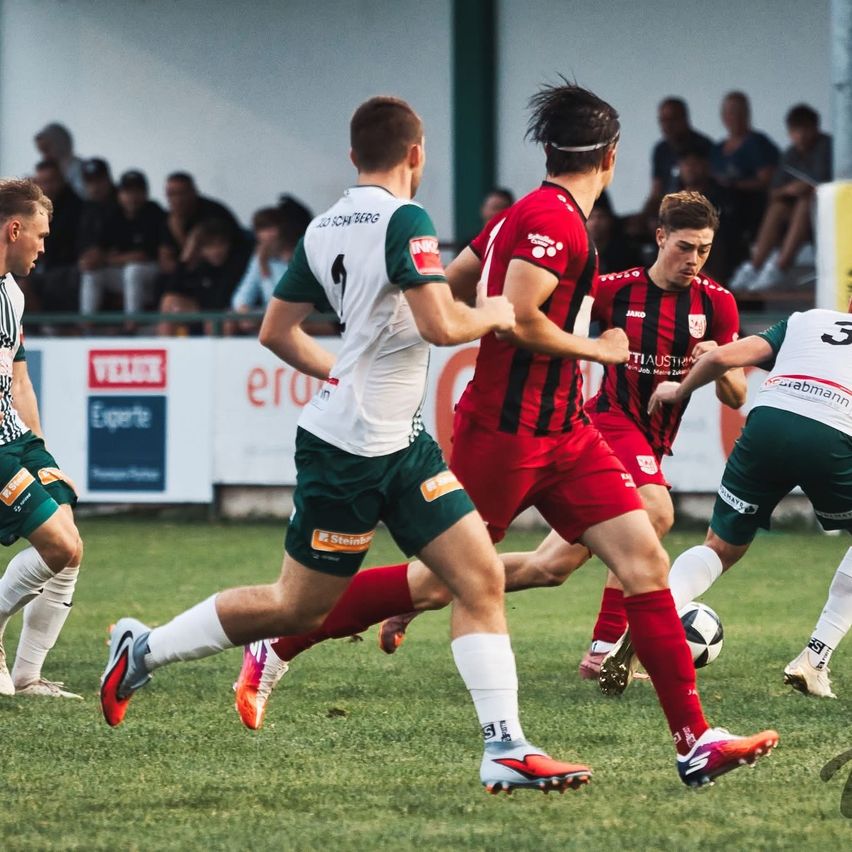 Fußballspieler in grünen und roten Trikots spielen auf einem Feld. Ein Spieler in Rot versucht, den Ball zu treten, während ein Spieler in Grün verteidigt.