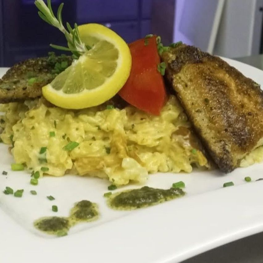 A plate of creamy pasta with a fried fish fillet, topped with a lemon slice and a red tomato.