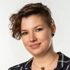 Eine Frau mit lockigem Haar und einer Kette mit braunem Anhänger lächelt für ein Porträt vor weißem Hintergrund. Sie trägt ein schwarz-weiß kariertes Hemd.
