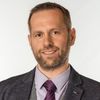 Ein Mann mit Bart lächelt und trägt einen grauen Anzug, ein lila Hemd und eine lila Krawatte.
