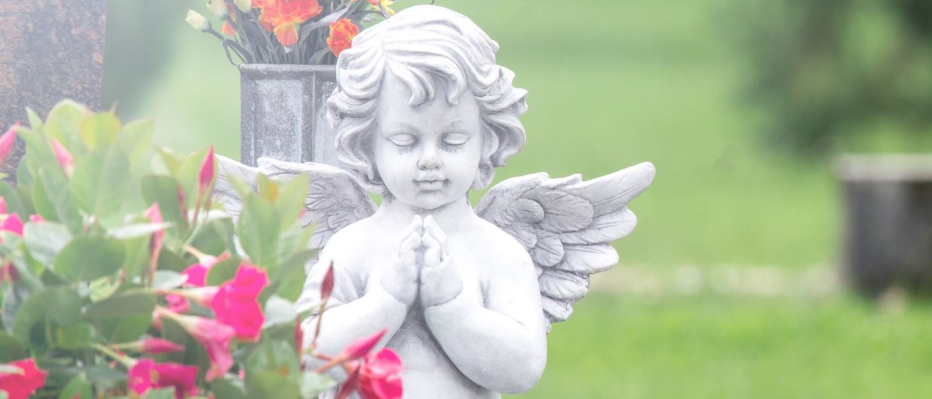 Eine weiße Engelsstatue steht auf einem Grab in einem Friedhof mit grünem Rasen und Blumen. Im Hintergrund befindet sich ein Grabstein.