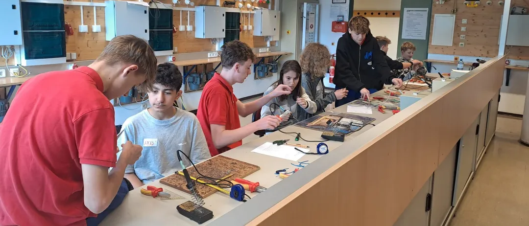 Eine Gruppe von Schülern in einem Klassenzimmer arbeitet an einem wissenschaftlichen Projekt. Sie verwenden Werkzeuge und Materialien auf einer Theke, mit einer Tafel und Postern im Hintergrund.