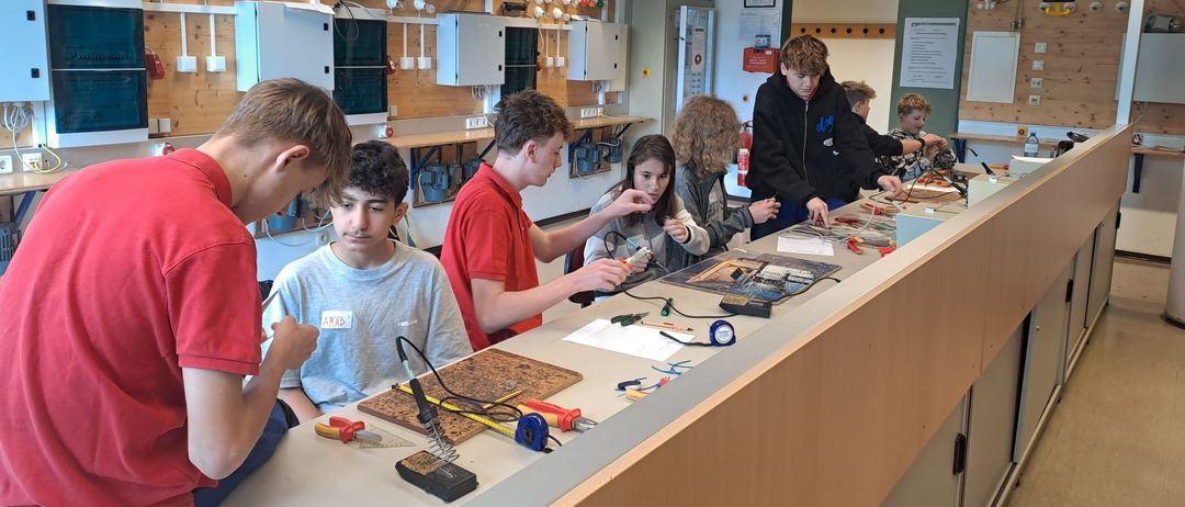 Eine Gruppe von Schülern in einem Klassenzimmer arbeitet an einem wissenschaftlichen Projekt. Sie verwenden Werkzeuge und Materialien auf einer Theke, mit einer Tafel und Postern im Hintergrund.