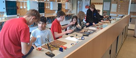 Eine Gruppe von Schülern in einem Klassenzimmer arbeitet an einem wissenschaftlichen Projekt. Sie verwenden Werkzeuge und Materialien auf einer Theke, mit einer Tafel und Postern im Hintergrund.