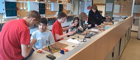 Eine Gruppe von Schülern arbeitet in einem Klassenzimmer mit einer Laborbank an einem Elektronikprojekt. Werkzeuge und Geräte sind auf der Bank verteilt. Der Raum hat eine Decke mit hängenden Objekten und Postern.