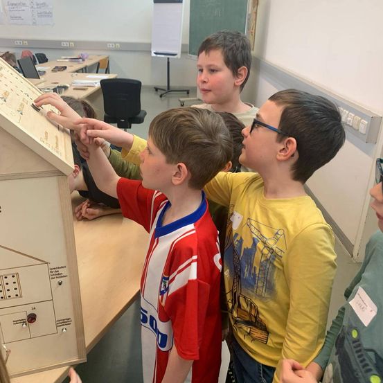 Vier Jungen in einem Klassenzimmer. Zwei von ihnen schauen auf ein Gerät auf einem Tisch. Das Gerät hat viele Schalter und Knöpfe.