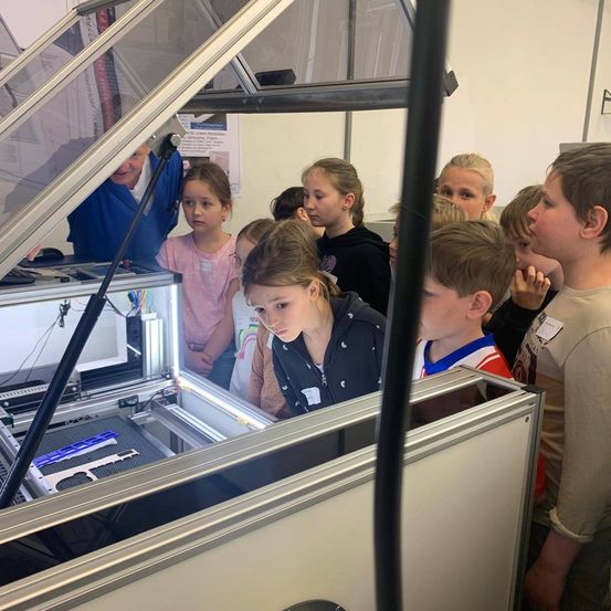 Kinder beobachten einen 3D-Drucker, der blaue Kunststoffteile in einem transparenten Behälter druckt.