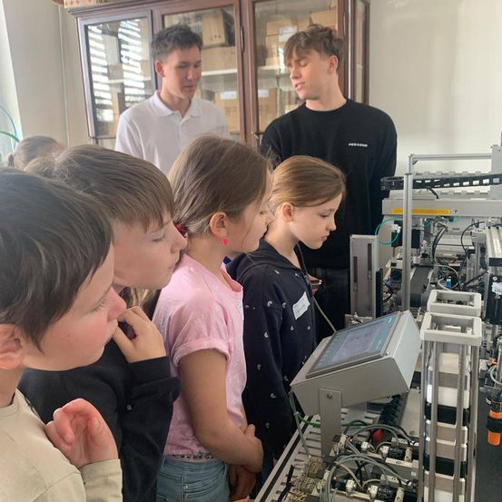 Eine Gruppe von Kindern beobachtet eine Maschine, während zwei Männer hinter ihnen stehen.