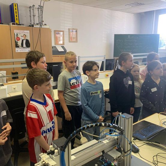 Eine Gruppe von Kindern versammelt sich um einen 3D-Drucker in einem Klassenzimmer, mit einer Tafel und Monitoren im Hintergrund.