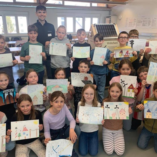 Klasse von Kindern in einem Klassenzimmer mit Solarmodulen und Windturbinen im Hintergrund, die Zeichnungen von Häusern und Windturbinen hochhalten.