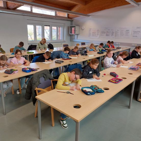 Schüler sitzen an Tischen in einem Klassenzimmer und arbeiten an ihren Aufgaben. Einige Schüler tragen eine Brille.