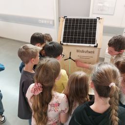 Eine Gruppe von Kindern versammelt sich um eine Ausstellung, die eine Windturbine zeigt. Die Ausstellung hat ein Solarpanel und Text über Windenergie.