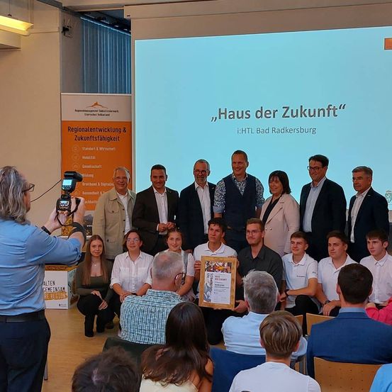 Eine Gruppe von Menschen in formeller Kleidung versammelt sich in einem Raum für eine Zeremonie. Sie posieren für ein Foto vor einer großen Leinwand.