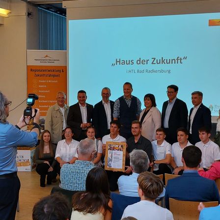 Eine Gruppe von Personen versammelt sich in einem Raum für eine Veranstaltung, mit einer Projektionsleinwand hinter ihnen, auf der 'Haus der Zukunft' steht. Sie posieren für ein Foto, einige halten Auszeichnungen. Eine Person links macht ein Foto mit einer Kamera.