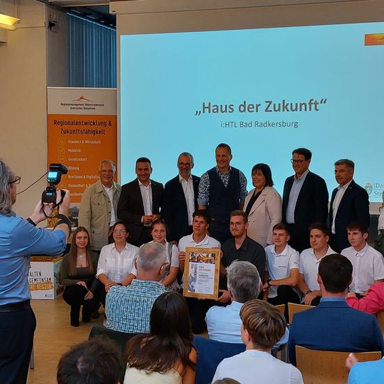 Eine Gruppe von Personen versammelt sich in einem Raum für eine Veranstaltung, mit einer Projektionsleinwand hinter ihnen, auf der 'Haus der Zukunft' steht. Sie posieren für ein Foto, einige halten Auszeichnungen. Eine Person links macht ein Foto mit einer Kamera.