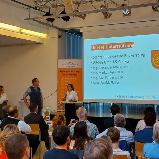 Eine Frau hält eine Präsentation an einem Podium in einem Konferenzsaal. Hinter ihr ist eine große Leinwand, die eine Folie mit dem Text 'Unsere Unterstützung' anzeigt. Mehrere Personen sitzen vor ihr. Auf der rechten Seite befindet sich ein Banner mit den Worten 'Regionalentwicklung & Zukunftsfähigkeit'.