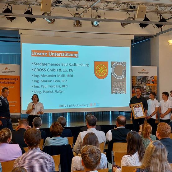 Eine Präsentation in einem Konferenzraum mit einem sitzenden Publikum. Eine Person steht an einem Podium mit einem Mikrofon. Eine Leinwand zeigt eine Liste von Sponsoren an. Eine Gruppe von Personen auf der Bühne hält einen Preis.