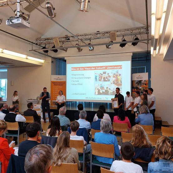 Eine Gruppe von Menschen versammelt sich in einem Raum, um eine Präsentation zu verfolgen. Die Präsentation wird auf einer Projektionsleinwand angezeigt. Die Menschen sitzen auf Stühlen, und einige stehen auf der Bühne.