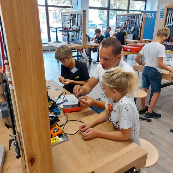 Ein Mann und zwei Kinder arbeiten in einer Werkstatt an einem Elektronikprojekt. Verschiedene Werkzeuge und Geräte stehen auf dem Tisch vor ihnen.