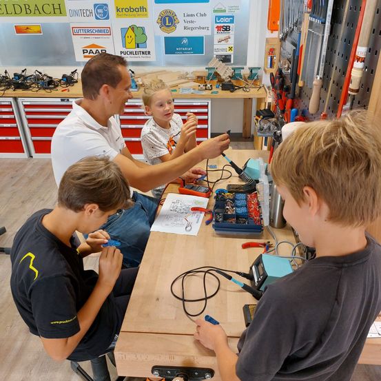 Ein lächelnder Mann instruiert drei junge Jungen in einer Werkstatt mit verschiedenen Werkzeugen und elektronischen Komponenten auf einem Tisch.