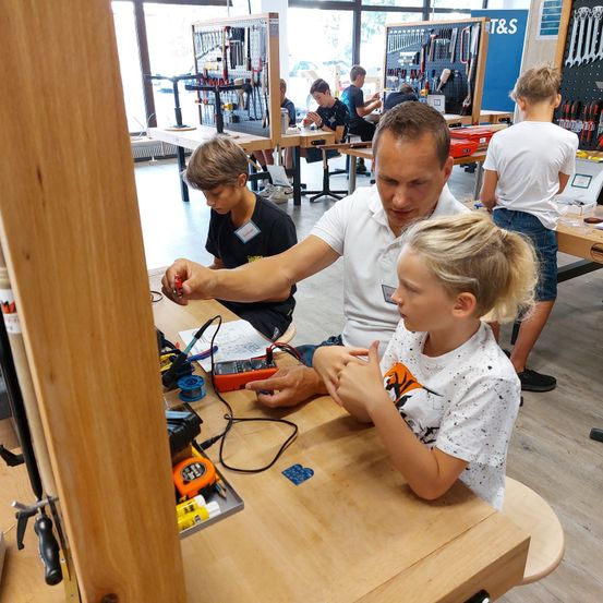 Ein Mann und zwei Kinder arbeiten in einer Werkstatt an einem Projekt. Sie sind von Werkzeugen und Ausrüstung umgeben. Der Mann demonstriert den Kindern etwas.