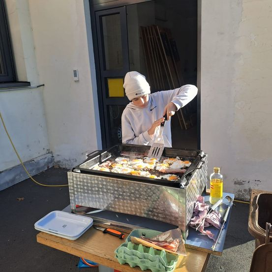 Eine Person kocht auf einem Outdoor-Grill mit mehreren Eiern und einem Pfannenwender. Die Szene spielt sich vor einem Gebäude ab.