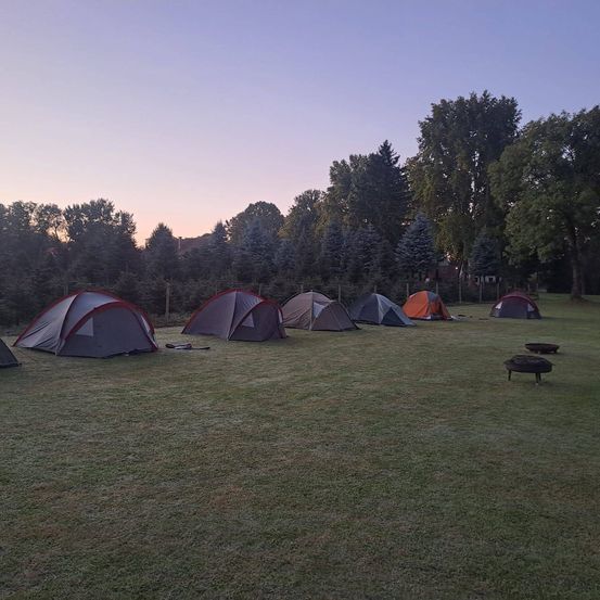 Eine Gruppe Zelte, die in einem Grasfeld mit Bäumen und Bergen im Hintergrund während des Sonnenuntergangs aufgebaut sind.