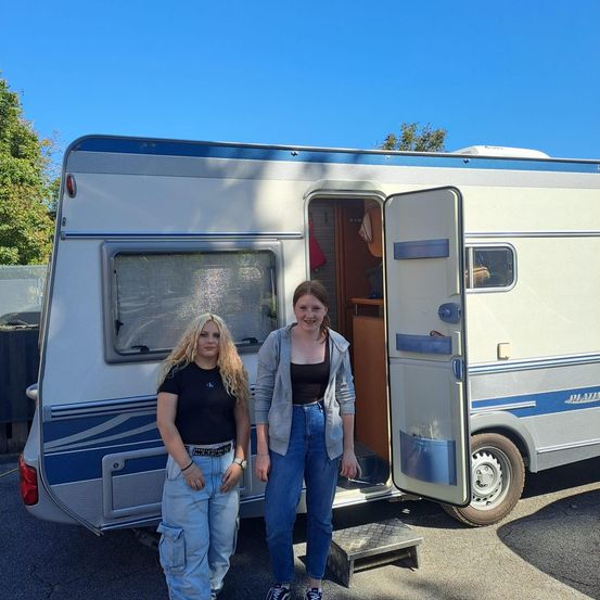 Zwei Frauen stehen vor einem offenen Wohnwagen und posieren lächelnd für ein Foto. Eine trägt ein schwarzes T-Shirt und Jeans, die andere eine graue Jacke und Jeans. Dahinter sind Bäume und ein klarer blauer Himmel zu sehen.