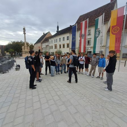 Eine Gruppe von Schülern, lässig gekleidet, versammelt sich auf einem Stadtplatz. Hinter ihnen wehen Flaggen verschiedener Länder an Masten. Links steht ein Denkmal, und im Hintergrund befinden sich Gebäude mit Türmen.