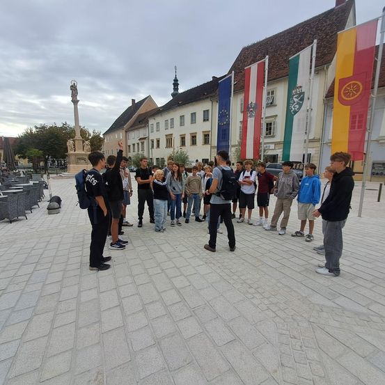 Eine Gruppe von Schülern, lässig gekleidet, versammelt sich auf einem Stadtplatz. Hinter ihnen wehen Flaggen verschiedener Länder an Masten. Links steht ein Denkmal, und im Hintergrund befinden sich Gebäude mit Türmen.