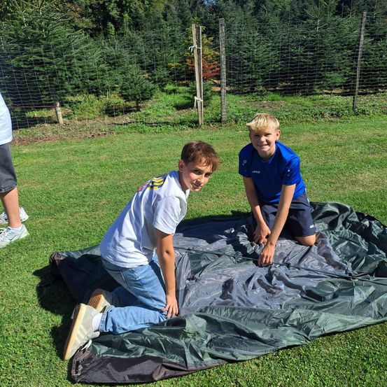 Zwei Jungen richten eine Tarp auf einem Grasfeld ein, mit einem Zaun und Bäumen im Hintergrund. Ein Junge kniet und der andere sitzt.