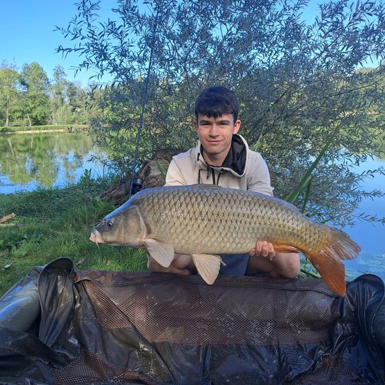 Ein junger Mann hält einen großen Karpfen in seinen Händen am See. Er lächelt für die Kamera.