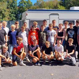 Eine Gruppe von Menschen, wahrscheinlich Schüler, posiert für ein Foto vor einem Wohnwagen, mit Bäumen und einem Zaun im Hintergrund.
