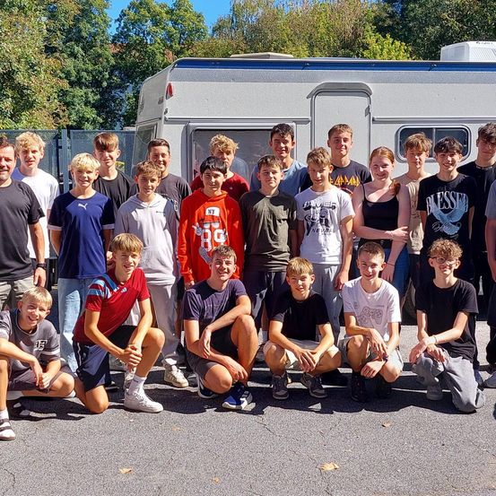 Eine Gruppe von Menschen, wahrscheinlich Schüler, posiert für ein Foto vor einem Wohnwagen, mit Bäumen und einem Zaun im Hintergrund.