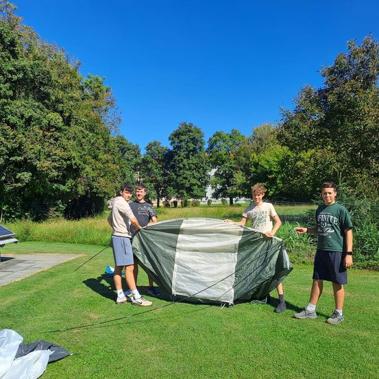 Vier Jungen stehen an einem sonnigen Tag in einem Park. Sie bauen ein grünes Zelt auf dem Rasen auf. Bäume und Pflanzen sind im Hintergrund.