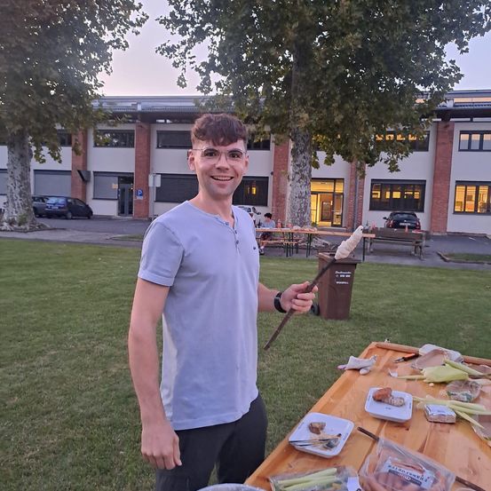 Ein Mann mit Brille und einem grauen T-Shirt hält einen Stock mit Marshmallows und posiert in einem Park. Dahinter befindet sich ein Gebäude mit mehreren Fenstern und vor dem Gebäude geparkte Autos.