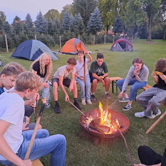 Eine Gruppe junger Leute versammelt sich um ein Lagerfeuer auf einer Rasenfläche. Sie sitzen auf Bänken und auf dem Boden und rösten Lebensmittel. Mehrere Zelte sind im Hintergrund zu sehen.