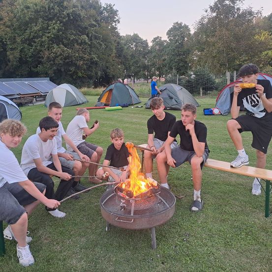 Eine Gruppe junger Jungen sitzt um ein Lagerfeuer in einem Campingbereich herum und röstet Essen. Bäume und Zelte sind im Hintergrund zu sehen.