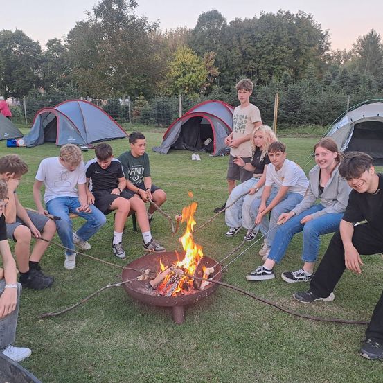 Eine Gruppe von Teenagern und Erwachsenen versammelt sich um ein Lagerfeuer, röstet Marshmallows, auf einer Wiese mit Zelten im Hintergrund.