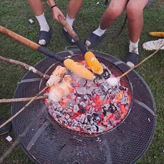 Zwei Personen grillen Maiskolben über einem Feuer auf dem Gras. Jede Person hält einen Stock mit Maiskolben. Der Grill ist mit brennenden Kohlen gefüllt.