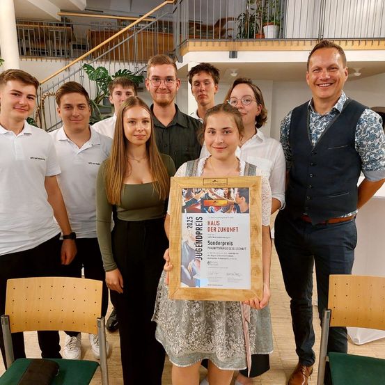 Eine Gruppe von Schülern und Erwachsenen steht lächelnd zusammen. Ein Mädchen hält einen gerahmten Zertifikat. Sie sind in einem Raum mit Holzstühlen und einer Treppe im Hintergrund.