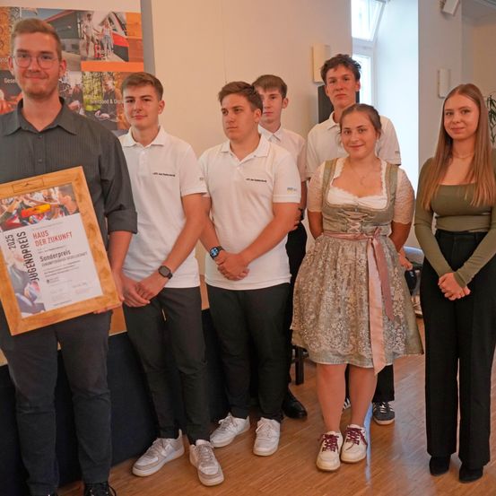 Eine Gruppe von Menschen steht drinnen und lächelt. Ein Mann hält einen gerahmten Preis. Sie tragen Freizeitkleidung, einige mit Turnschuhen. Ein Poster mit einem Baum ist im Hintergrund.