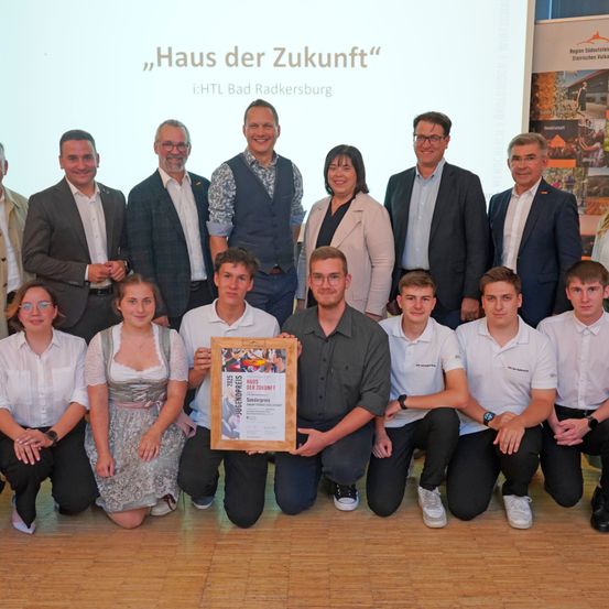 Eine Gruppe von Personen posiert für ein Foto, wobei ein Mann in der Mitte ein gerahmtes Zertifikat hält. Hinter ihnen zeigt eine Leinwand den Text 'Haus der Zukunft'. Sie sind in formeller Kleidung gekleidet, und einige tragen Brillen. Der Ort scheint ein Präsentationsort oder eine Preisverleihung zu sein.