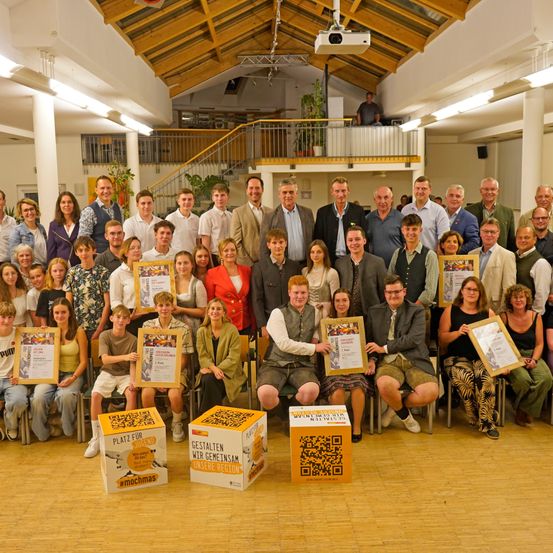 Eine Gruppe von Menschen posiert für ein Foto in einem Saal. Einige von ihnen halten Auszeichnungen. Boxen mit Text stehen vor ihnen. Eine Treppe befindet sich hinter ihnen. Lampen sind an der Decke montiert. Eine Person steht auf dem Balkon. Ein Projektor hängt von der Decke.