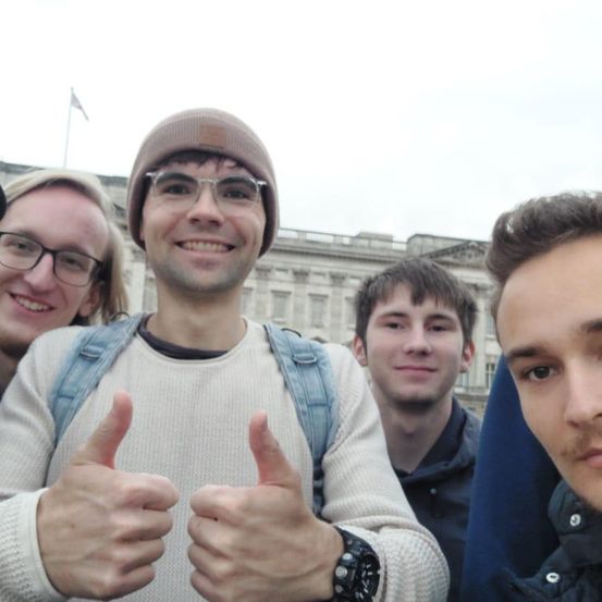 Fünf junge Männer, die lächeln und Daumen hoch halten, stehen vor einem historischen Gebäude, möglicherweise einem Palast. Sie tragen Freizeitkleidung.