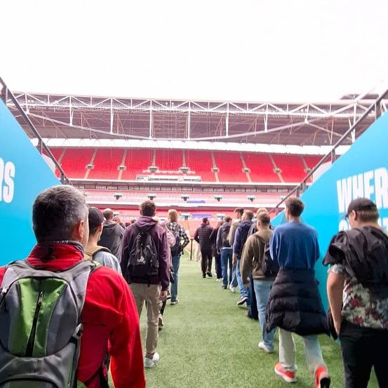 Eine Gruppe von Menschen geht auf dem Spielfeld eines großen Stadions mit leeren roten Sitzen und blauen Bannern entlang.