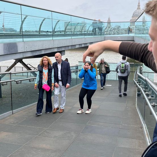 Ein Paar und eine Frau gehen auf einer Glasbrücke mit einem Fluss und Gebäuden im Hintergrund. Ein anderer Mann macht ein Foto.