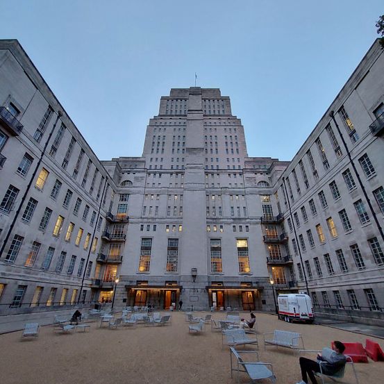 Ein Außenhof vor einem großen Gebäude mit vielen Fenstern. Stühle und Tische sind im Hof aufgestellt, und Menschen sitzen und stehen. Das Gebäude ist aus Stein und hat ein modernes architektonisches Design.